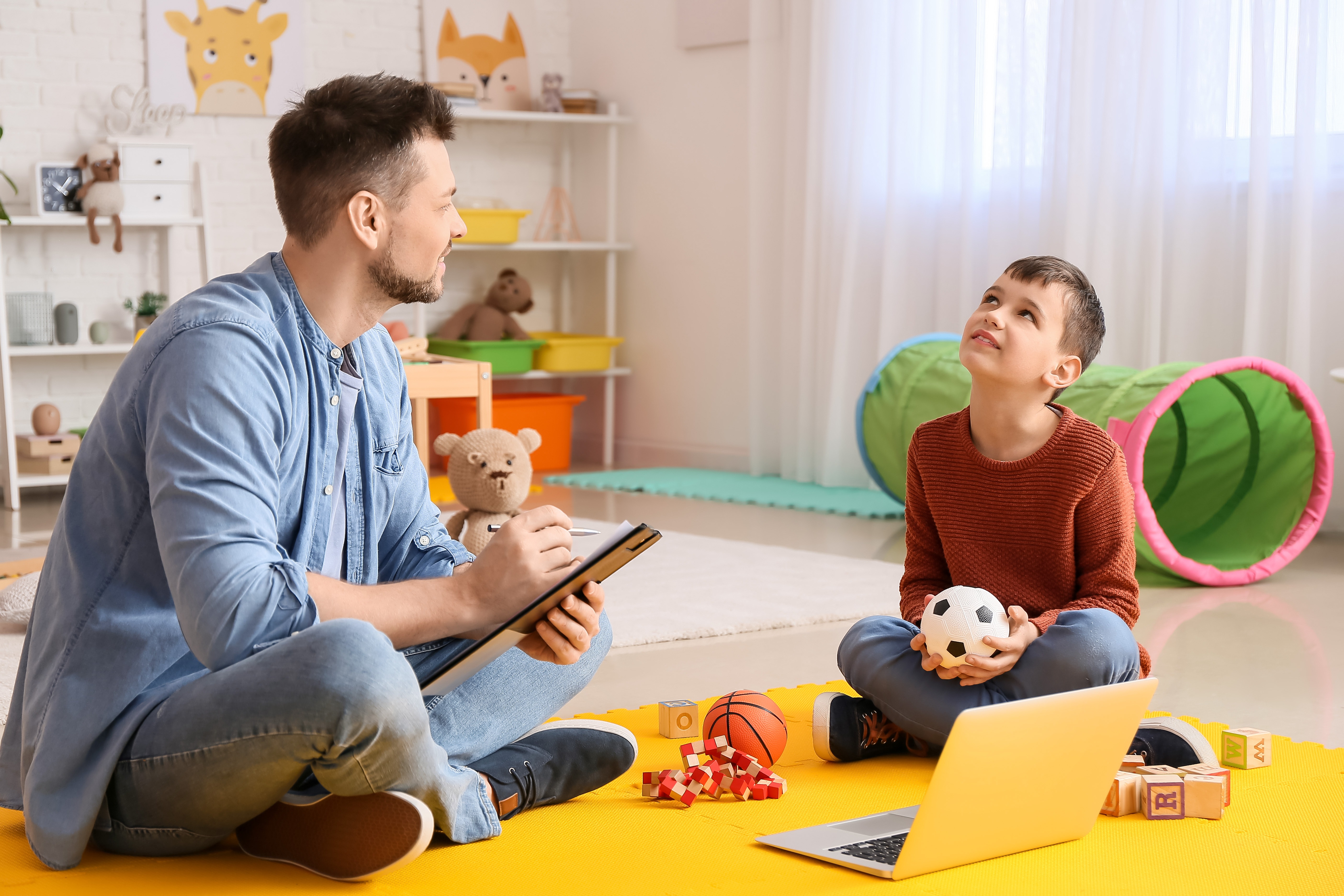 ABA therapist taking notes while a boy holding a soccer ball sits on a play mat
